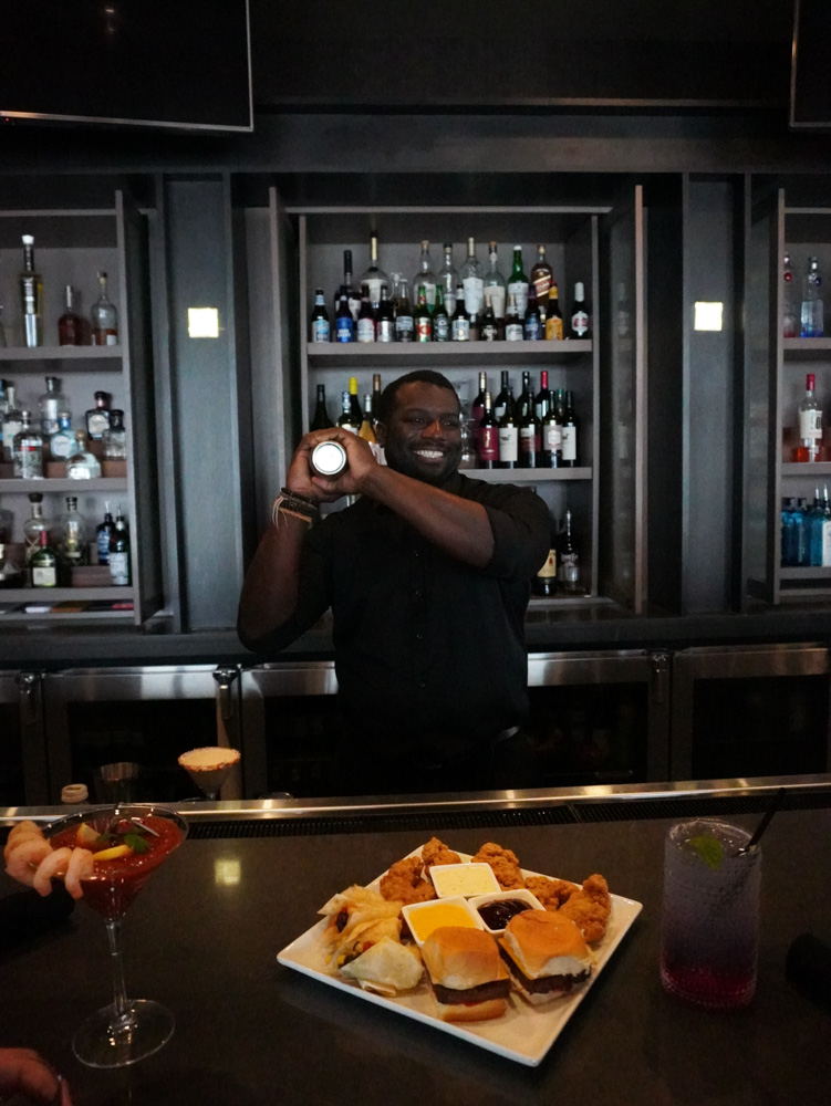 A bartender standing behind a bar, holding a cocktail shaker. He is wearing a black shirt and has a tray of food in front of him.