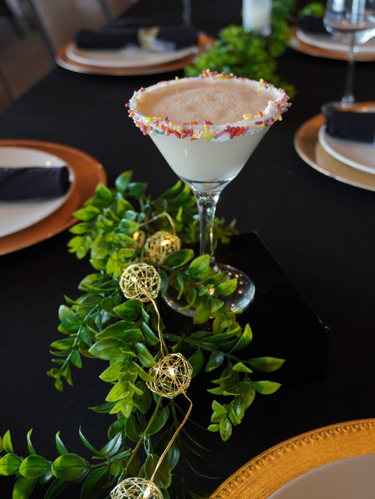 A white cocktail in a martini glass with a sprinkled rim, garnished with a green leafy branch and two golden balls.
